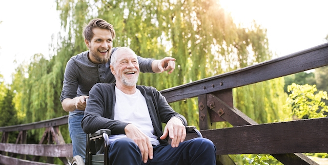 Young hipster son walking with disabled father in wheelchair on wooden bridge at park, pointing finger at something. Carer assisting disabled senior man.